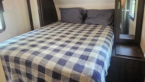 A bedroom with a checkered bedspread, a dark headboard, a mirror, and a wooden floor. - Wildwood camper #5  at Heaven's Ridge Campground (Fair Play)