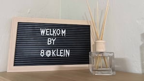 Un cartello di benvenuto incorniciato con la scritta 'WELCOME BY 8@KLEIN' e una bottiglia di vetro trasparente con bastoncini di canna accanto.