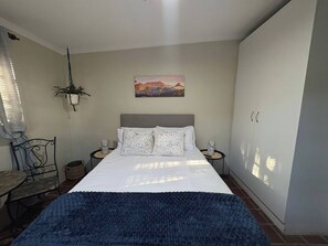 Un dormitorio con cama, mesita de noche, lámpara, planta en maceta y un cuadro en la pared.