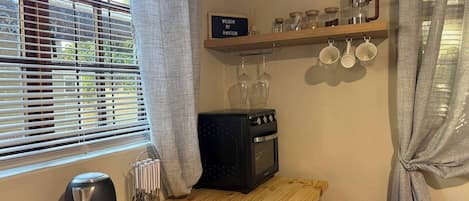 A kitchen with a stove, kettle, and a wooden shelf with cups and jars.
