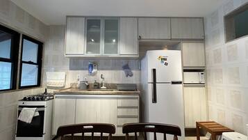 Een functionele keuken met een gasfornuis, een koelkast en een magnetron. Er staan twee houten stoelen en een klein krukje.