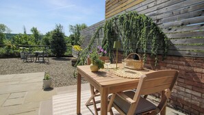 Eine Terrasse im Freien mit einem Holztisch und Stühlen, einem gewebten Korb, einem Blumentopf und einer Wand mit Kletterpflanzen.