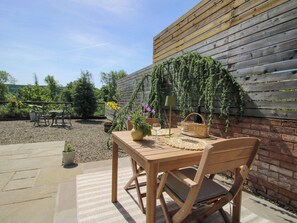 An outdoor patio with a wooden table and chairs, a woven basket, a potted plant, and a wall with climbing plants. - Thyme Away (Bucknell)