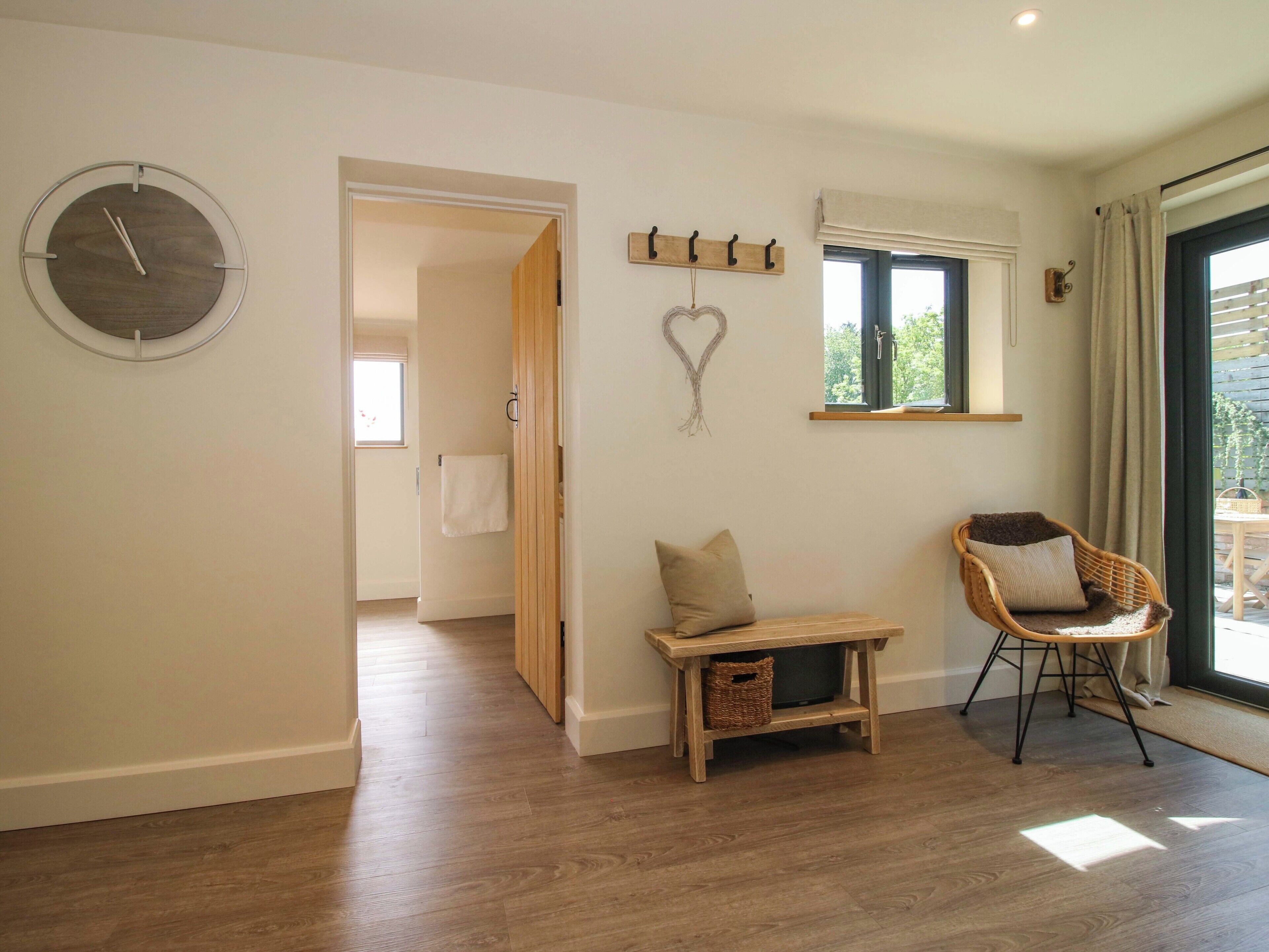 A room with a wooden floor, a round wall clock, a small table with a cushion, a wicker chair, and a heart-shaped decoration.