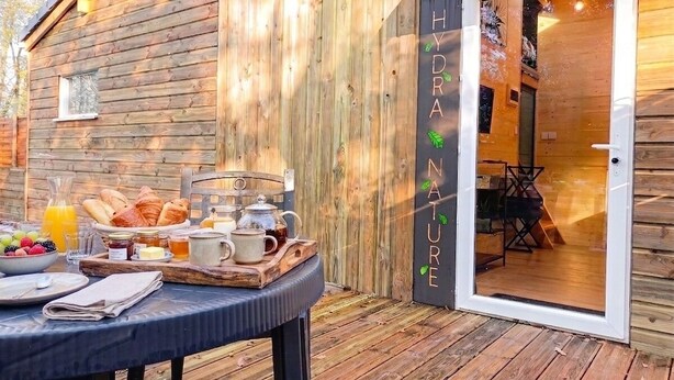 A wooden cabin with a breakfast setup on the deck, a door with 'HYDRAN NATURE' signage, and a glimpse into the interior. - Gîte Hydra Nature (Conteville)