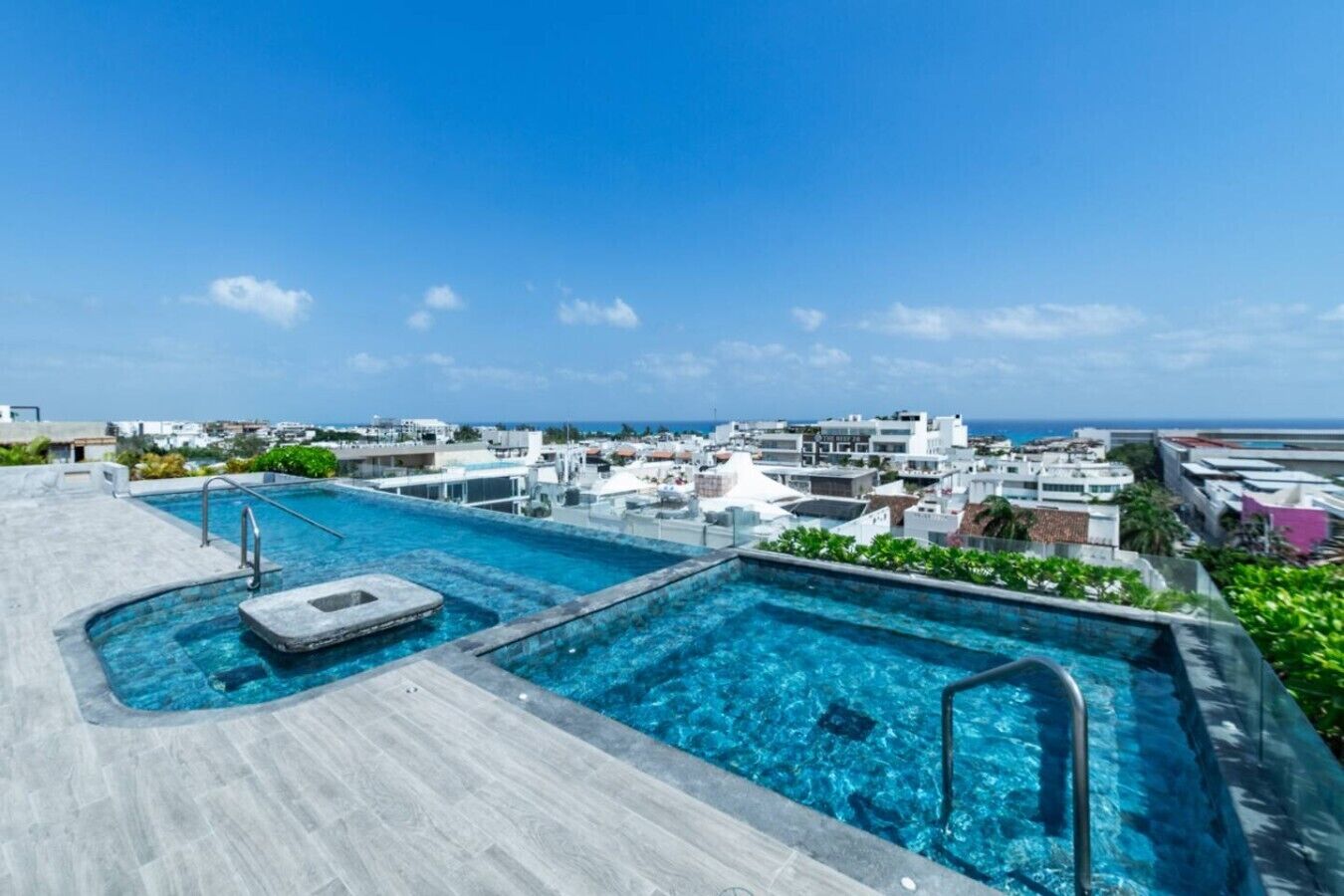 A rooftop pool with clear blue water, surrounded by a wooden deck and a view of the cityscape and ocean.