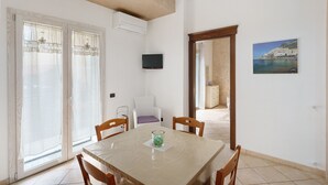 A dining area with a table, chairs, a TV, and a view of a coastal town through a window. - Casa Anna Standard Faito View in Pimonte – Comfort e Vista Montagna (Pimonte)