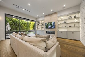 Ein modernes Wohnzimmer mit einer Eckcouch, einem an der Wand montierten Fernseher und einem großen Fenster mit Blick auf einen Garten.