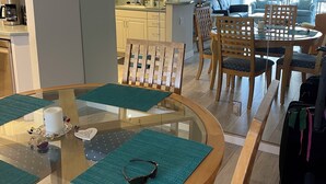 Moderni olohuone, jossa on lasinen ruokapöytä, puiset tuolit, keittiöalue ja näkymä ulos.