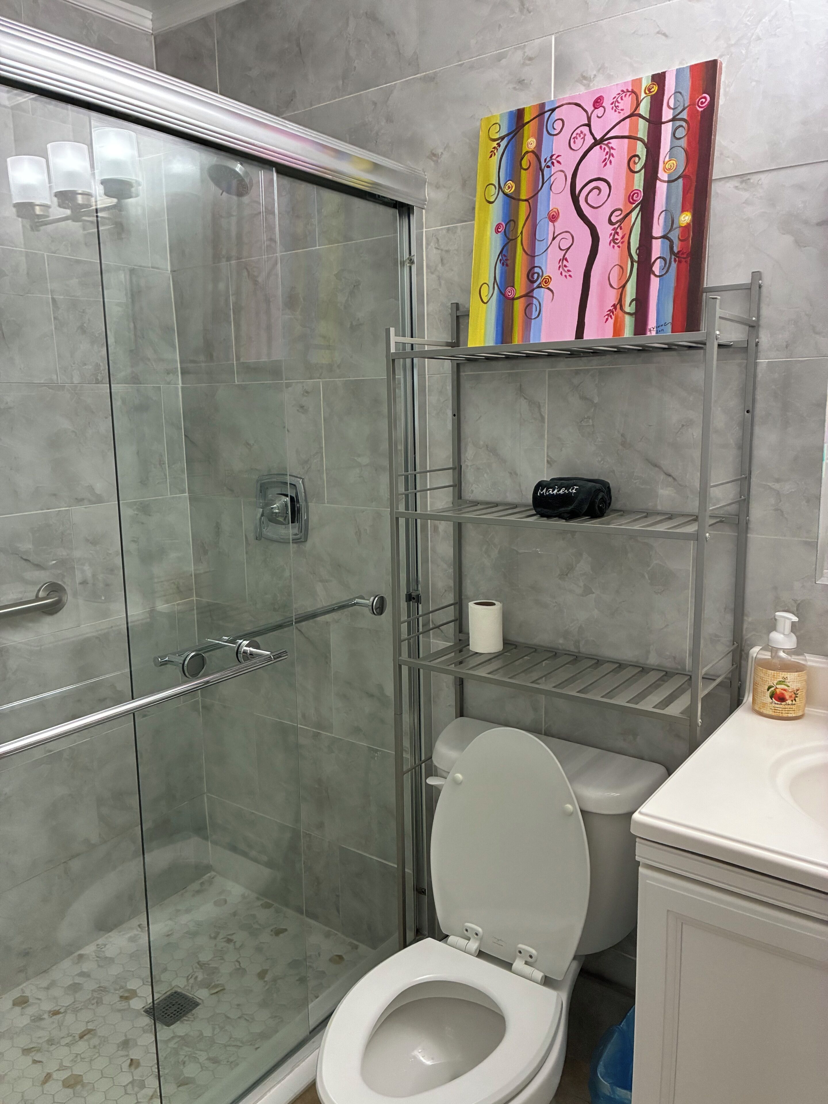 Un baño con cabina de ducha de vidrio, un inodoro, un estante con artículos de aseo personal y una obra de arte colorida en la pared.