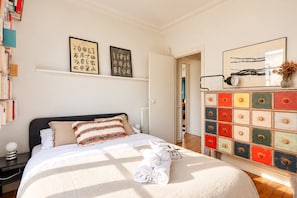 A neatly made bed with white linens and rolled towels, a colorful dresser with multiple drawers, and two framed botanical prints on the wall. - Saint Germain - 3BR - Notre-Dame - BnBNova (Paris)