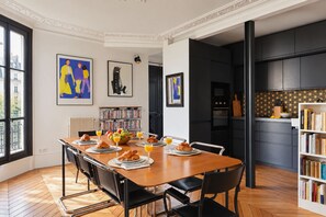 A modern kitchen with a dining area, featuring a wooden table set for breakfast, a bookshelf, and artwork on the walls. - Saint Germain - 3BR - Notre-Dame - BnBNova (Paris)