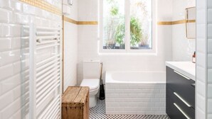 Un baño con paredes de azulejos blancos, una ventana con vista a follaje, una bañera blanca y un taburete de madera.