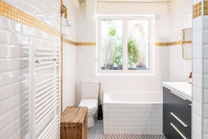 Ein Badezimmer mit weißen Fliesenwänden, einem Fenster mit Blick auf Grünflächen, einer weißen Badewanne und einem hölzernen Hocker.