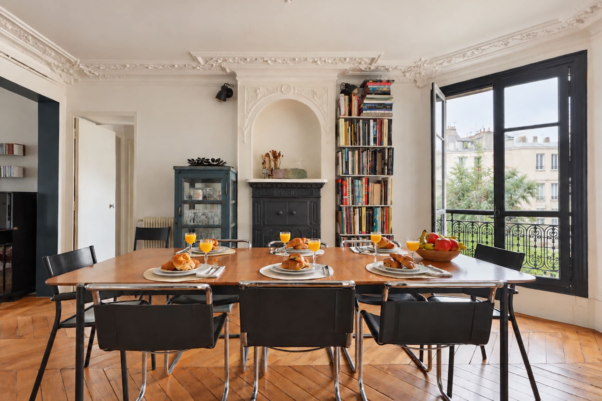 A dining area with a wooden table set for breakfast, surrounded by black chairs, with a bookshelf and a fireplace in the background.