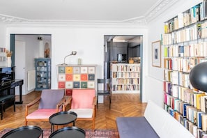 Ein Wohnzimmer mit einem Klavier, einer Bücherwand, einem Sessel, einer Couch und einem Fischgrätparkettboden.