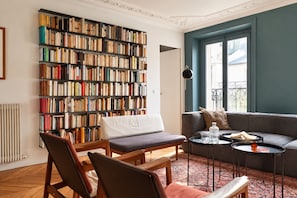 A cozy living room with a large bookshelf, a sofa, a coffee table, and a view of the outside. - Saint Germain - 3BR - Notre-Dame - BnBNova (Paris)