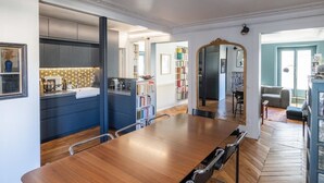 Una habitación amplia con una gran mesa de comedor de madera, una cocina con armarios oscuros y una estantería llena de libros.