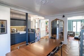 Ein geräumiges Zimmer mit einem großen Esstisch aus Holz, einer Küche mit dunklen Schränken und einer Bücherwand voller Bücher.