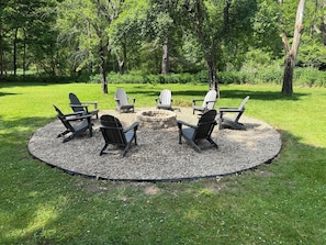 A circular seating area with Adirondack chairs around a stone fire pit, surrounded by gravel and grass, with trees in the background.