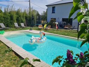 Ein Schwimmbecken mit einer Schwane als Schwimmpfau, umgeben von einer Holzterrasse, Stühlen und einem Haus im Hintergrund.