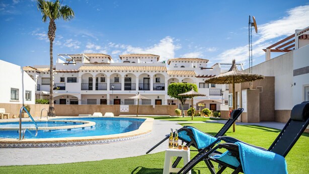Uma área de piscina com espreguiçadeiras, uma pequena mesa com bebidas e um edifício com varandas ao fundo.