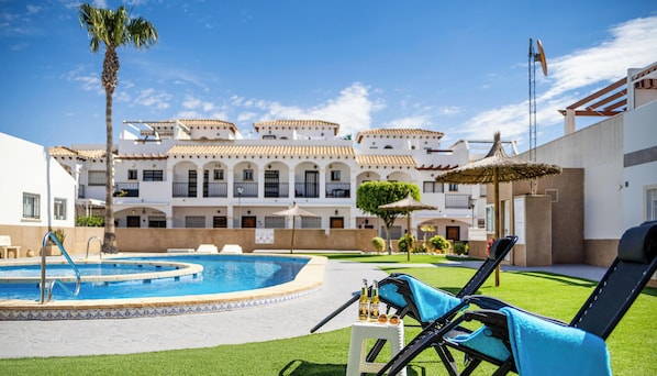 A pool area with lounge chairs, a small table with drinks, and a building with balconies in the background. - Sol y Brisa Apartment terrace swimming pool and BBQ Orihuela Costa (Orihuela)