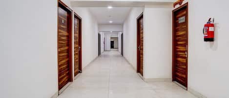 A long, straight hallway with wooden doors on both sides, a fire extinguisher on the right wall, and a ceiling light.