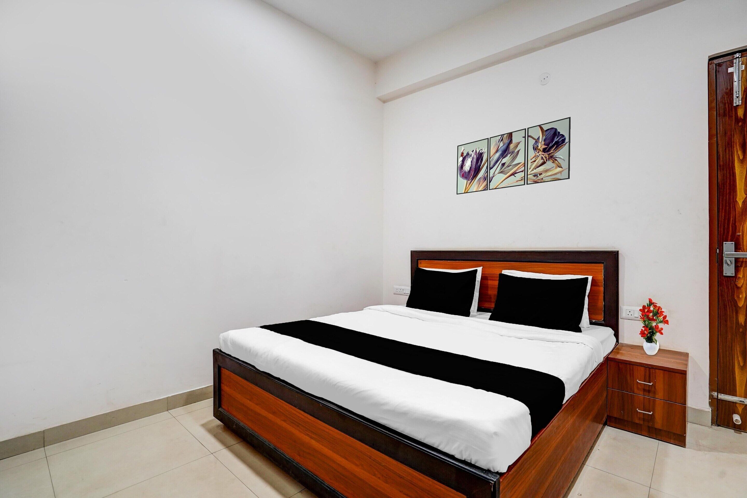 A bedroom with a wooden bed, white bedding, a nightstand with a vase, and a door with a wooden frame.