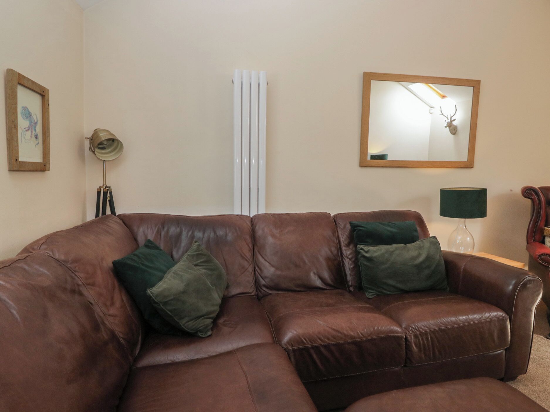 A brown leather sofa with two green cushions, a framed mirror with a deer head, a lamp, and a radiator.