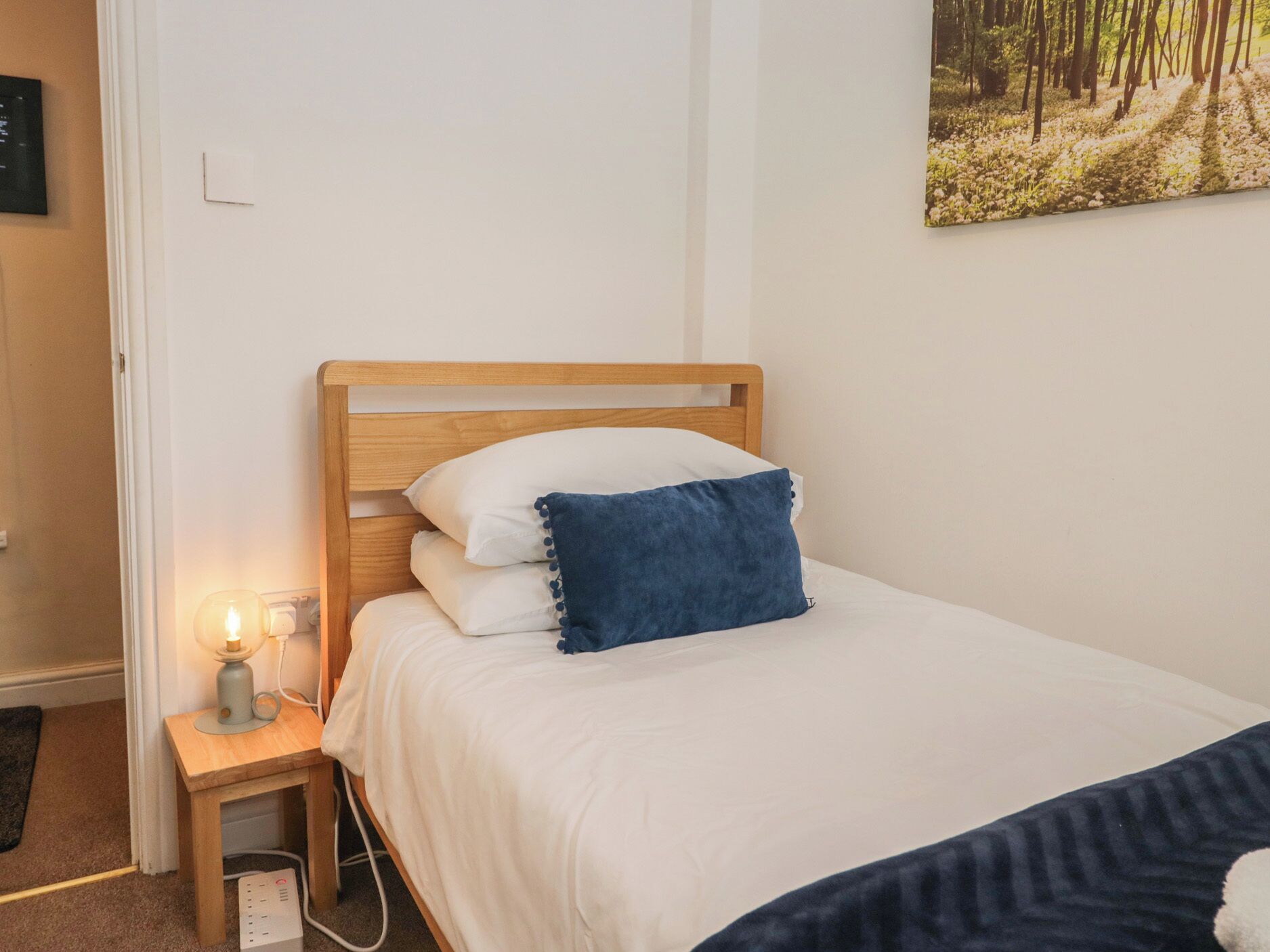 A bed with a wooden headboard, white bedding, a blue pillow, a bedside lamp, and a power strip.