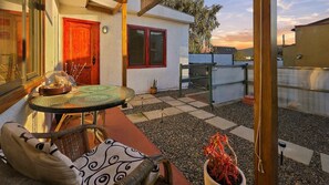 Un patio con mesa y sillas, un pequeño jardín y vistas al atardecer.
