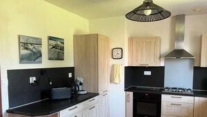 Una cocina moderna con armarios de madera clara, encimera negra, horno empotrado y cocina a gas. Hay dos relojes de pared y una lámpara colgante.