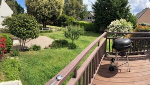Una terraza de madera con una parrilla, rodeada por un jardín vallado con diversas plantas y árboles.