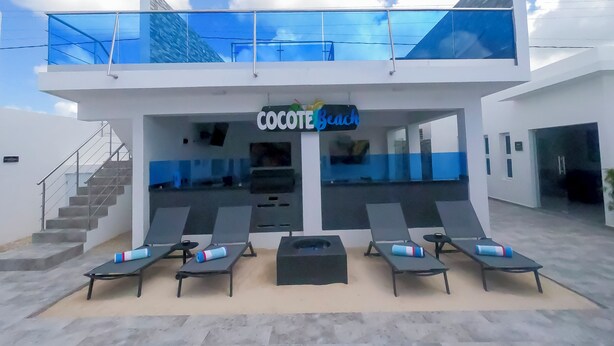 Un edificio moderno con un cartello che indica 'Cocote Beach' e un'area piscina con lettini e una scalinata.