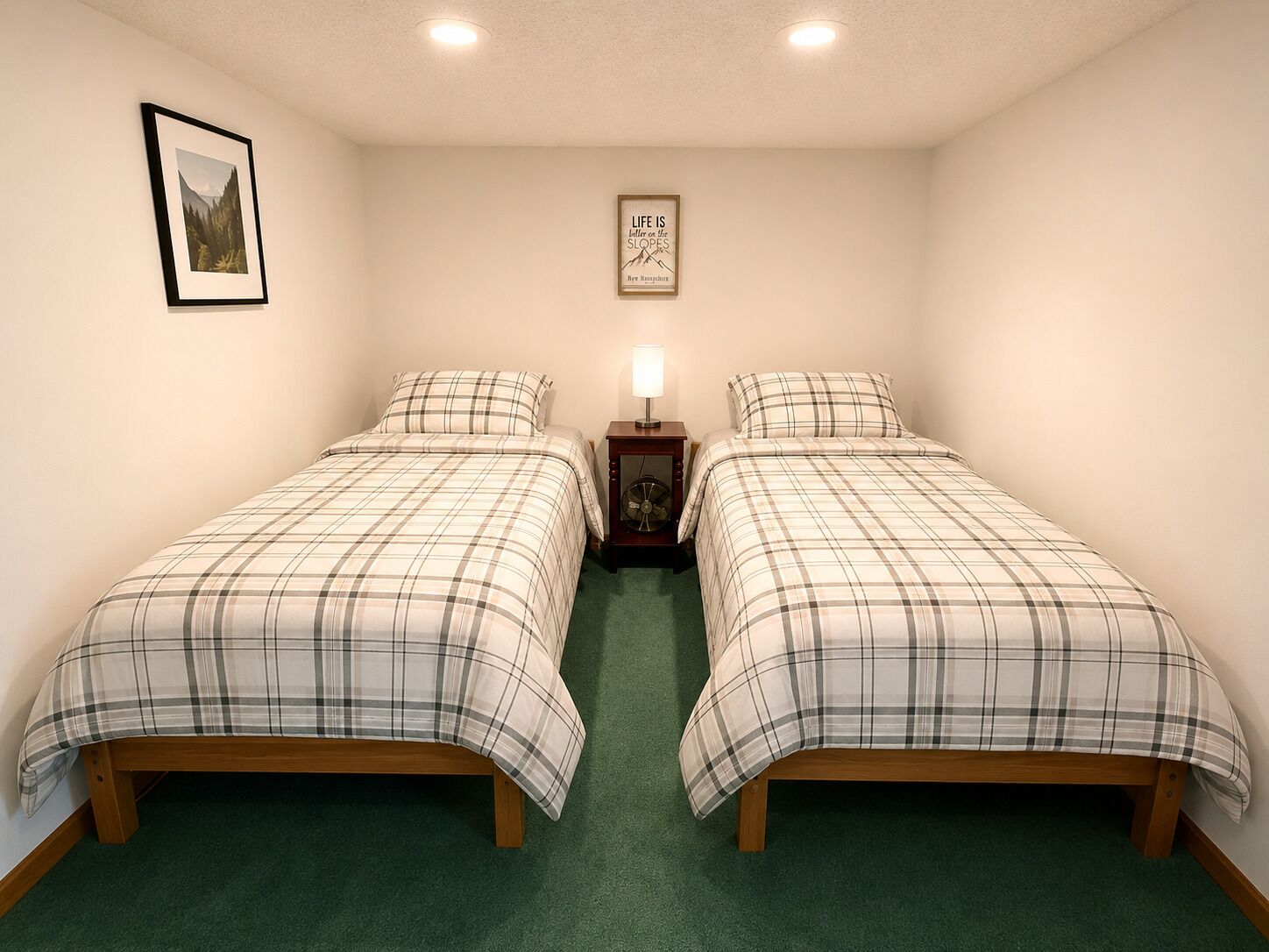 Ein Doppelzimmer mit zwei Betten, einem kleinen Nachttisch mit Lampe, einem gerahmten Zitat an der Wand und einem gerahmten Bild über dem Bett links.