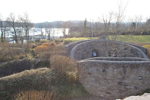 A stone structure with a small window, surrounded by vegetation and a body of water in the background.