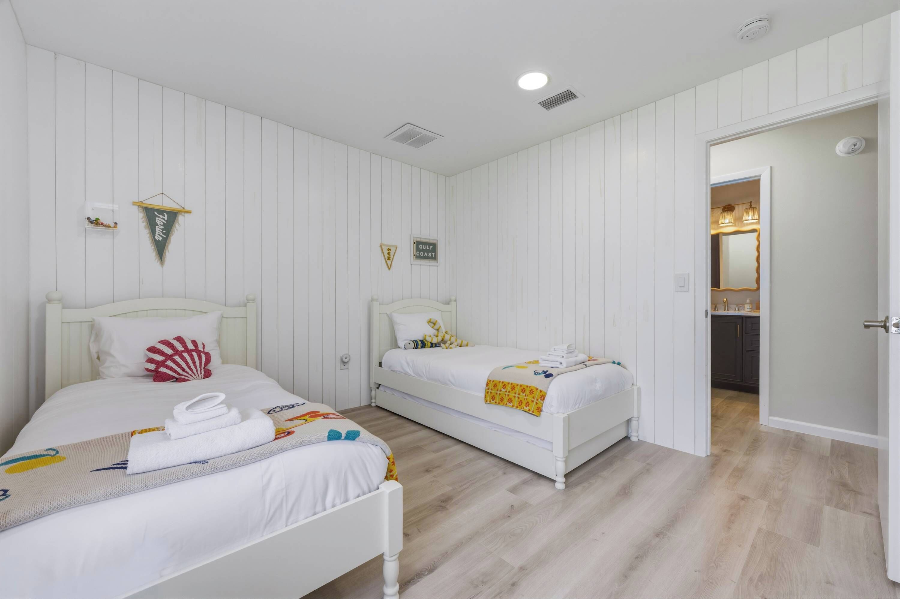 Habitación con dos camas, paredes blancas, suelo de madera, un estante pequeño con objetos decorativos y una puerta que da a otra habitación.