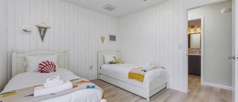 Habitación con dos camas, paredes blancas, suelo de madera, un estante pequeño con objetos decorativos y una puerta que da a otra habitación.