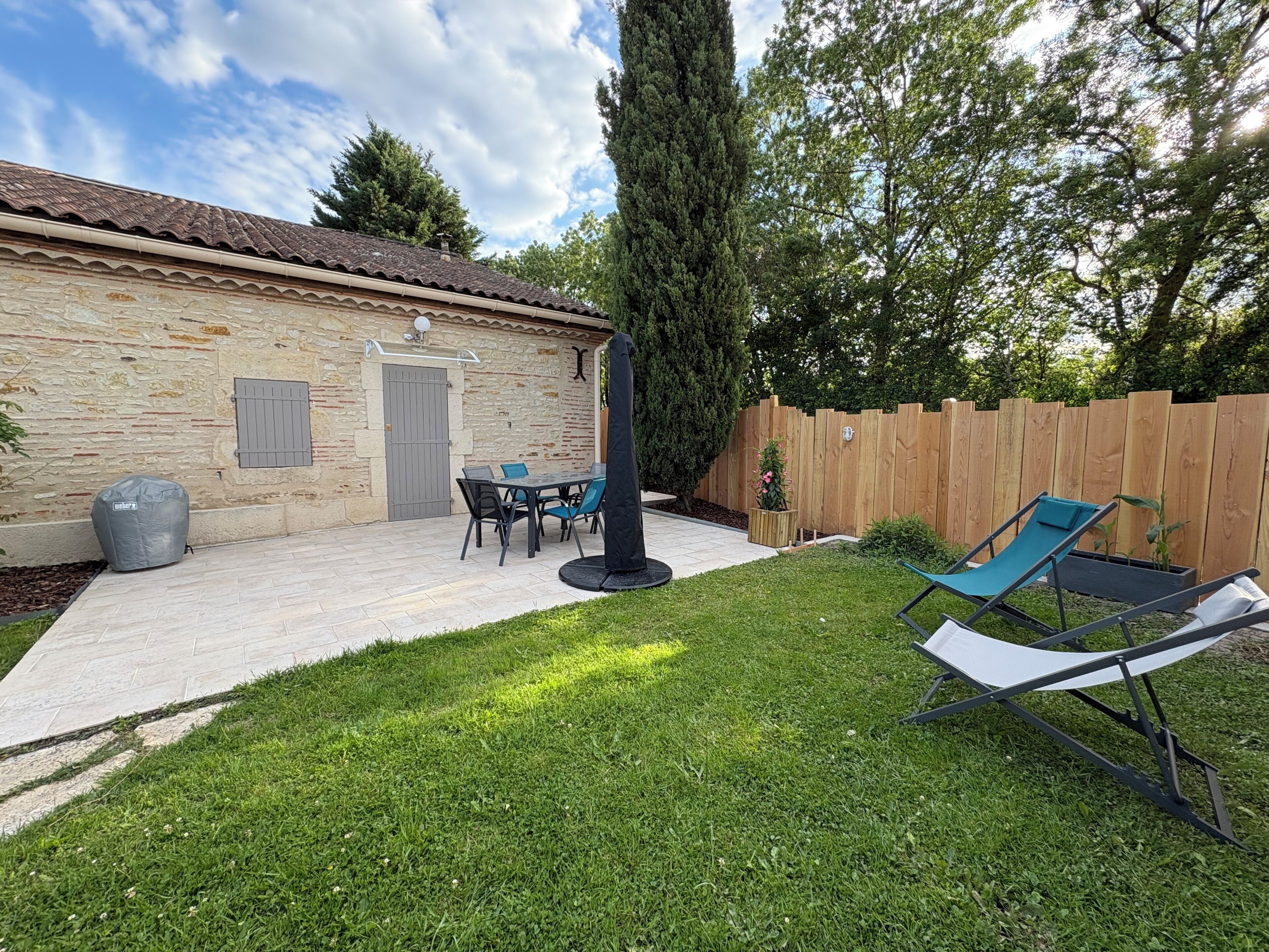 A stone building with a tiled roof, a patio with a table and chairs, a covered grill, and a wooden fence.