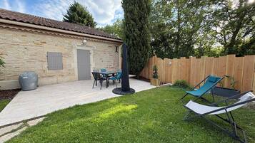 A stone building with a tiled roof, a patio with a table and chairs, a covered grill, and a wooden fence.