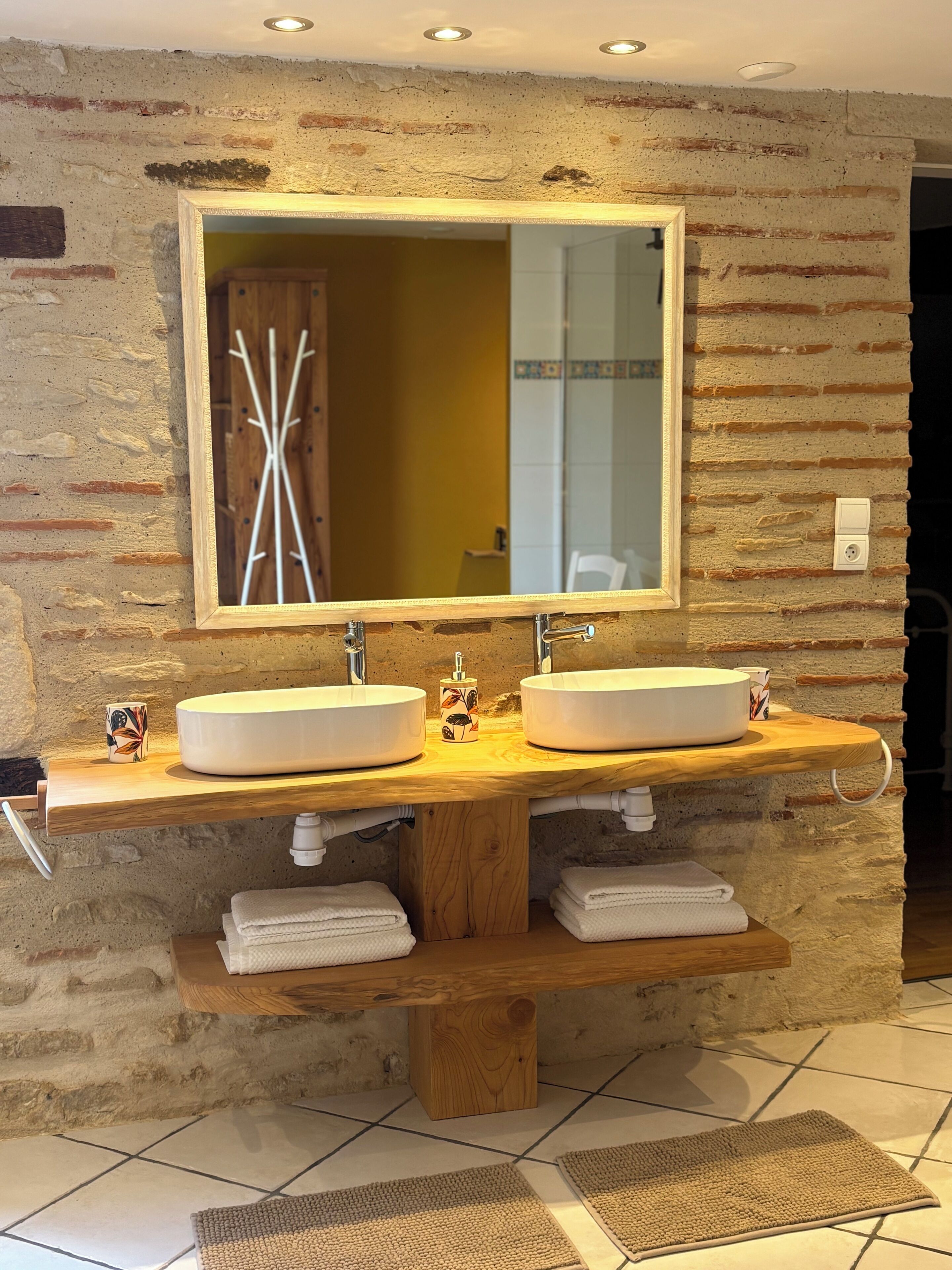 A bathroom with a rustic brick wall, a wooden vanity with two sinks, a large mirror, and a towel rack.