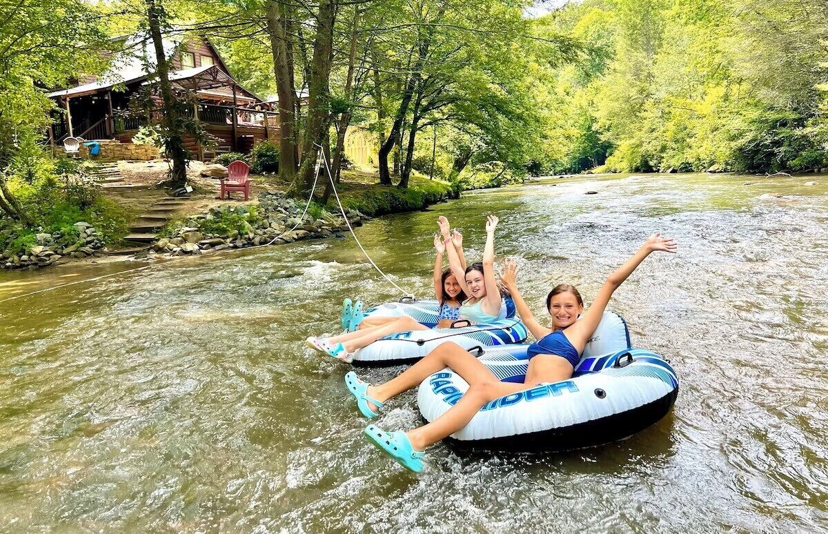 Two people on inflatable tubes in a river surrounded by trees and a cabin.