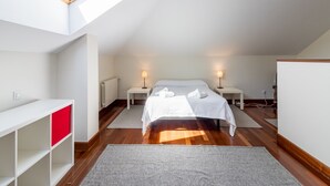 Un dormitorio con tragaluz, cama con ropa blanca, dos lámparas de noche, una consola blanca y un cajón rojo.