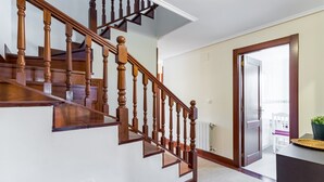 Una escalera de madera con barandilla, una pared blanca, una puerta que da a un balcón y una mesita con una planta.