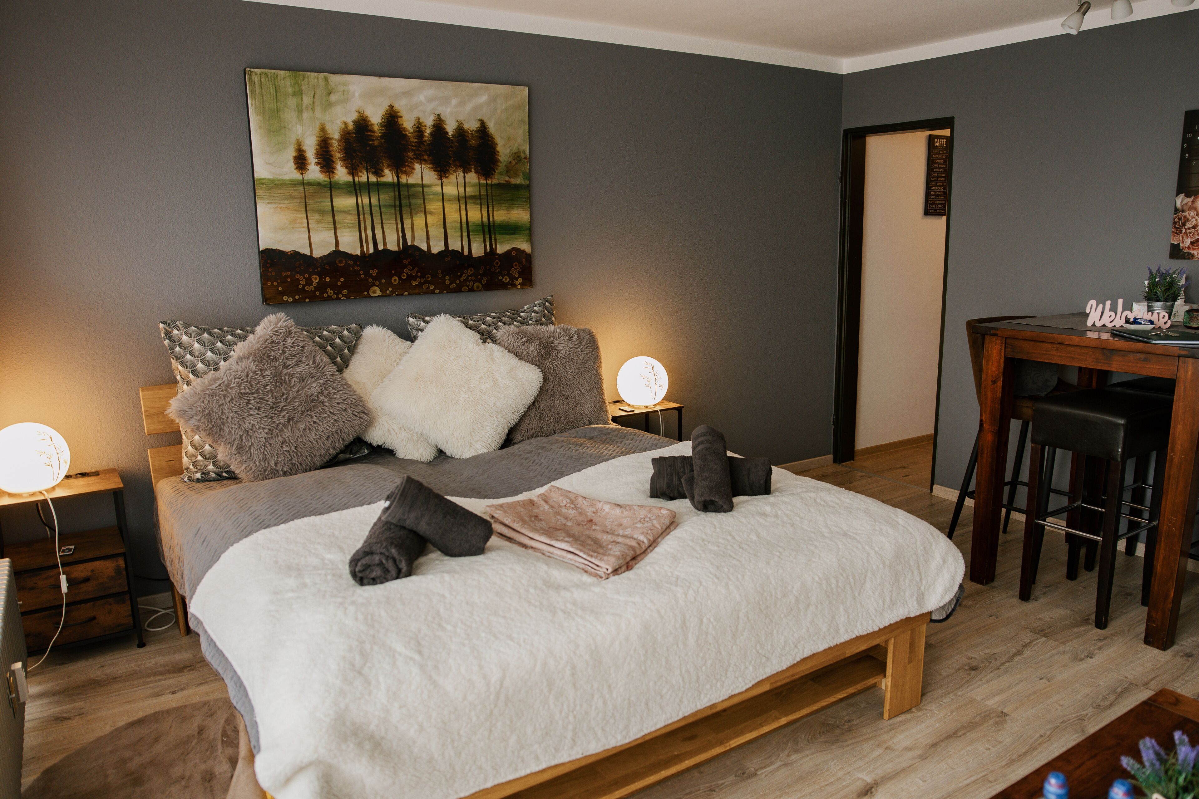Un dormitorio con una cama, almohadas, mesitas de noche, un cuadro y un escritorio con silla.