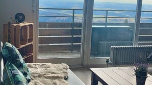 Une pièce dotée d’une grande fenêtre donnant sur la côte, d’un banc en bois, d’un tapis moelleux et d’une petite table sur laquelle est posée une plante.