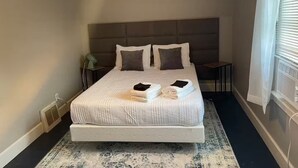 Un letto rifatto con cuscini, un comodino, una lampada e una finestra con persiane.