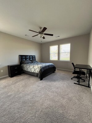 A bedroom with a bed, a desk, a chair, a ceiling fan, and two windows.