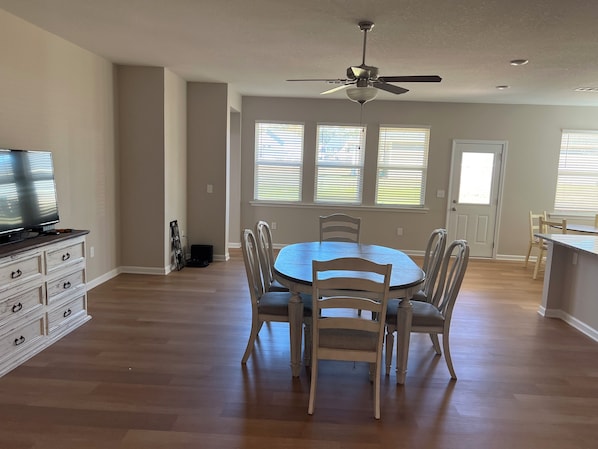 Un coin repas avec une table ronde, six chaises, un ventilateur de plafond et un téléviseur posé sur une commode.
